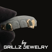 Gray dental grill with gold accents held by a gloved hand, branded 'by Grillz Jewelry'.